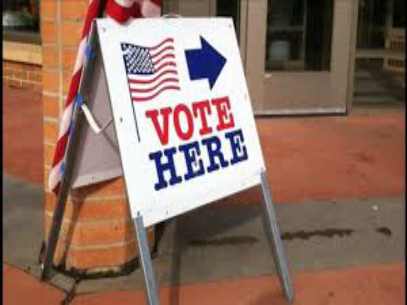A standing fold up sign angled away to the right towards the entrance of a building in the background. The sign has an American flag blowing to the right, an arrow pointing right